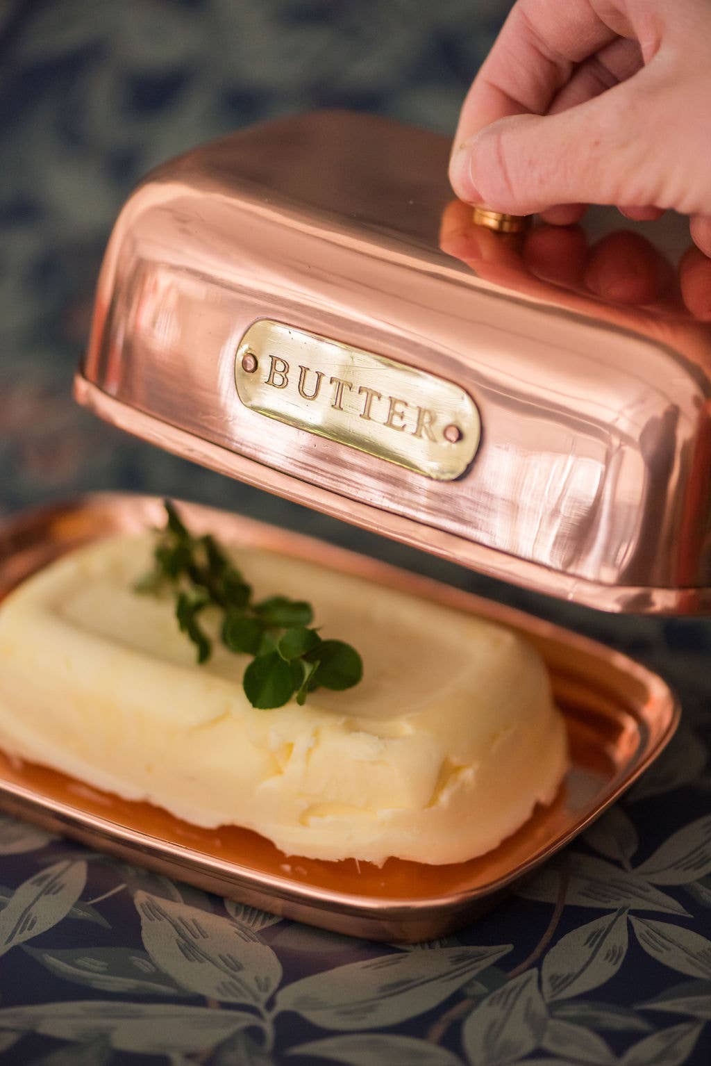 Copper butter dish with a pat of butter and a hand lifting the lid.