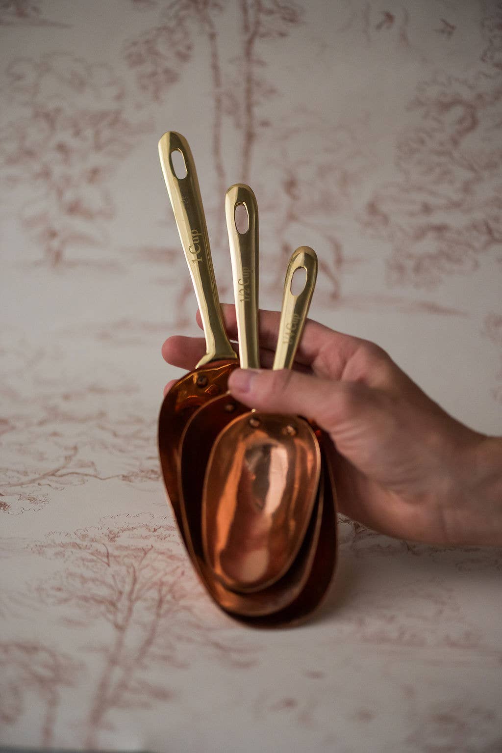 Hand holding a set of copper measuring spoons against a patterned background