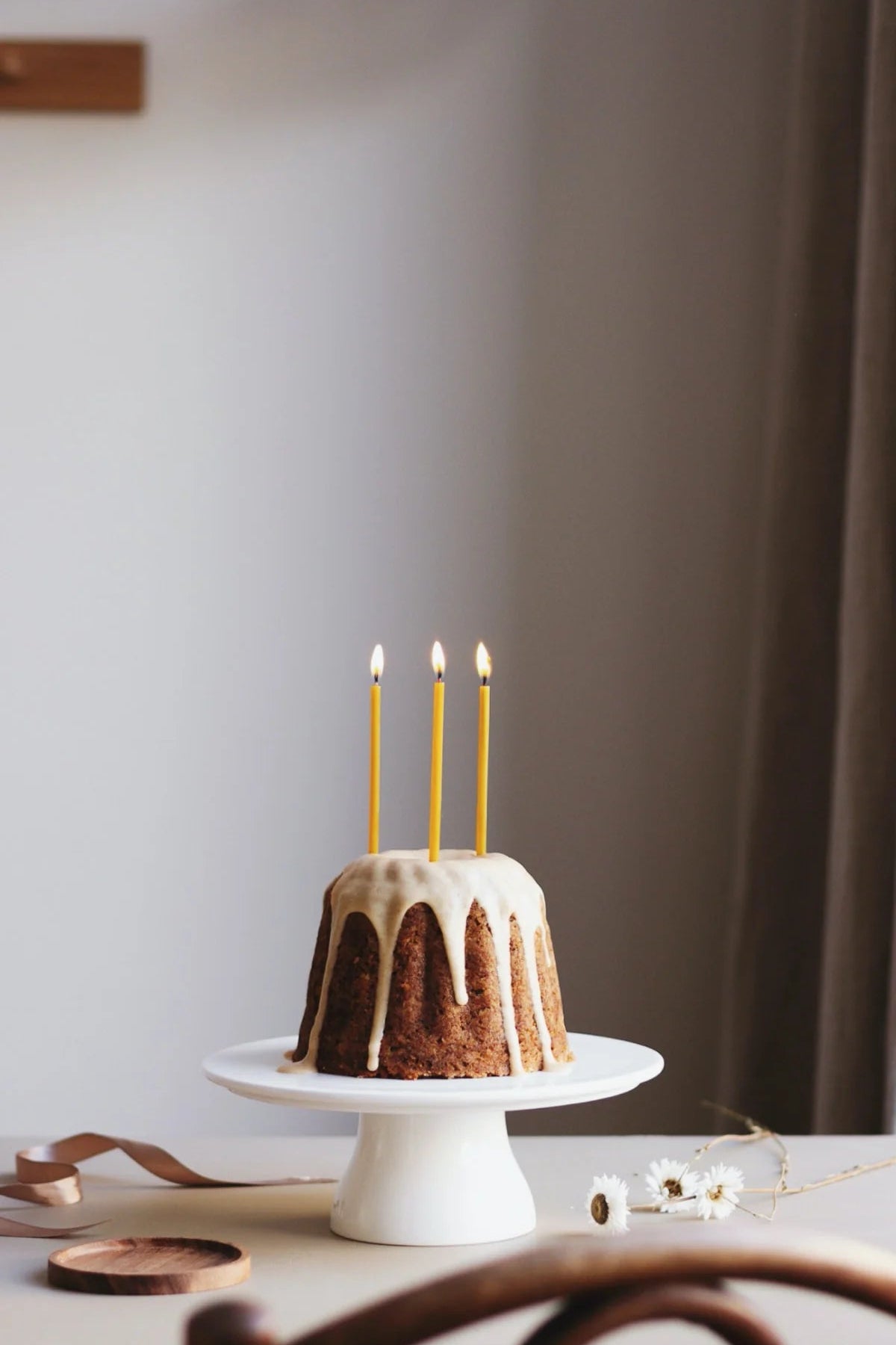 Small bundt cake with three beeswax birthday candles on a white cake stand against a plain wall.