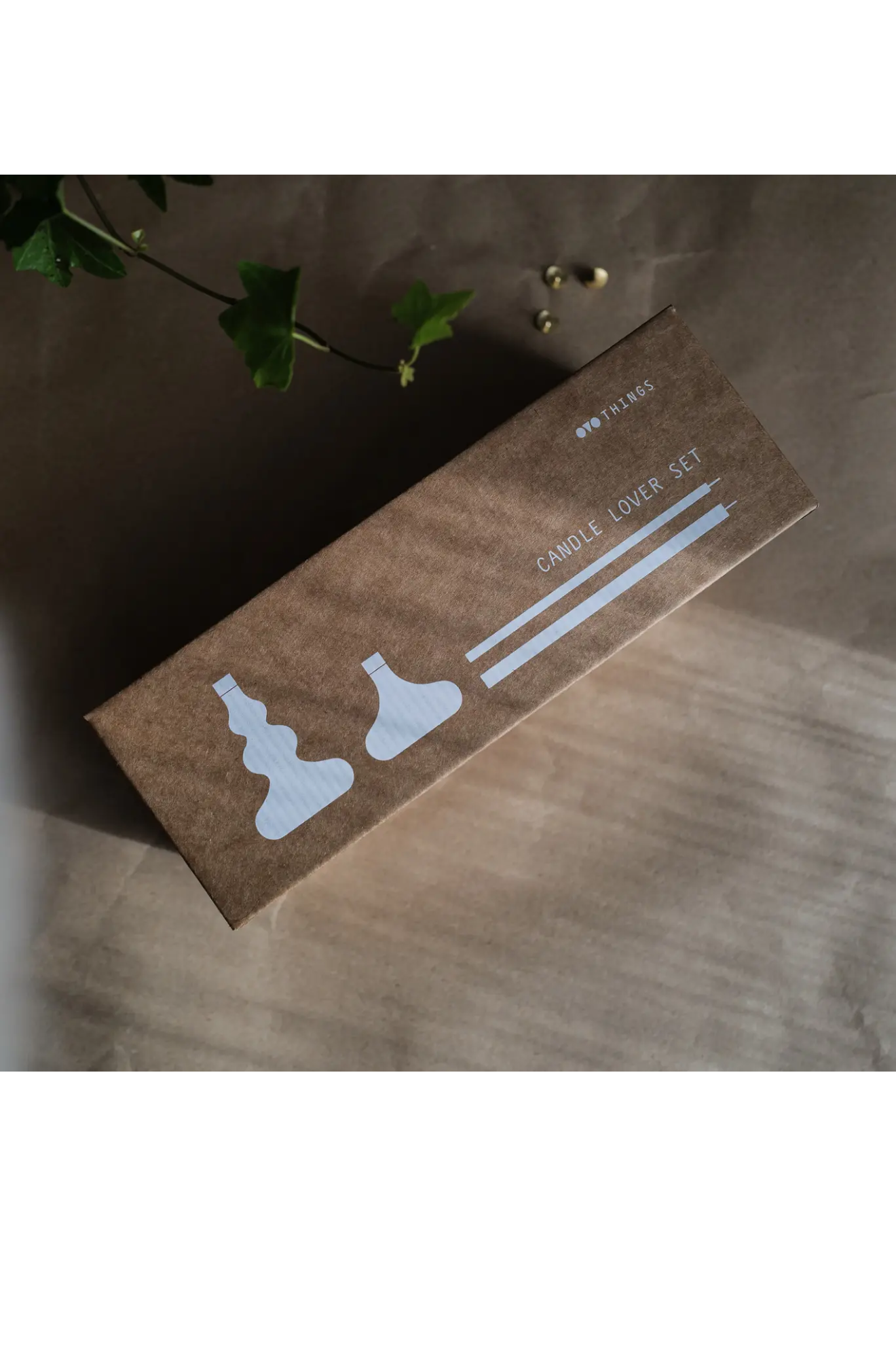 Box of candles and two candle holders on a wooden surface with a plant in the background