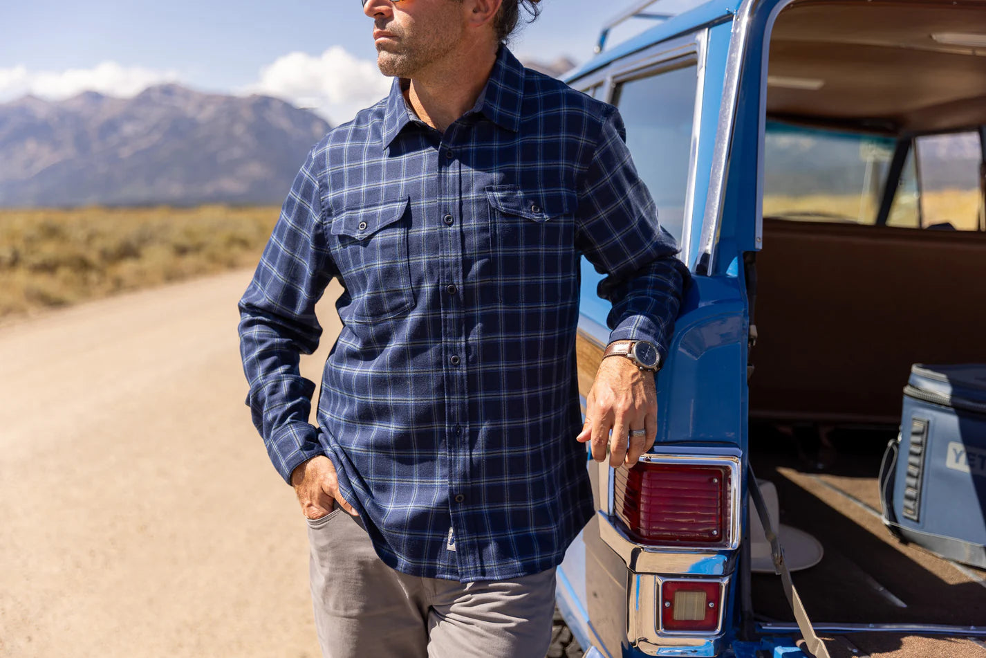 Man in a blue plaid shirt standing next to an open van in a desert setting with mountains in the background.