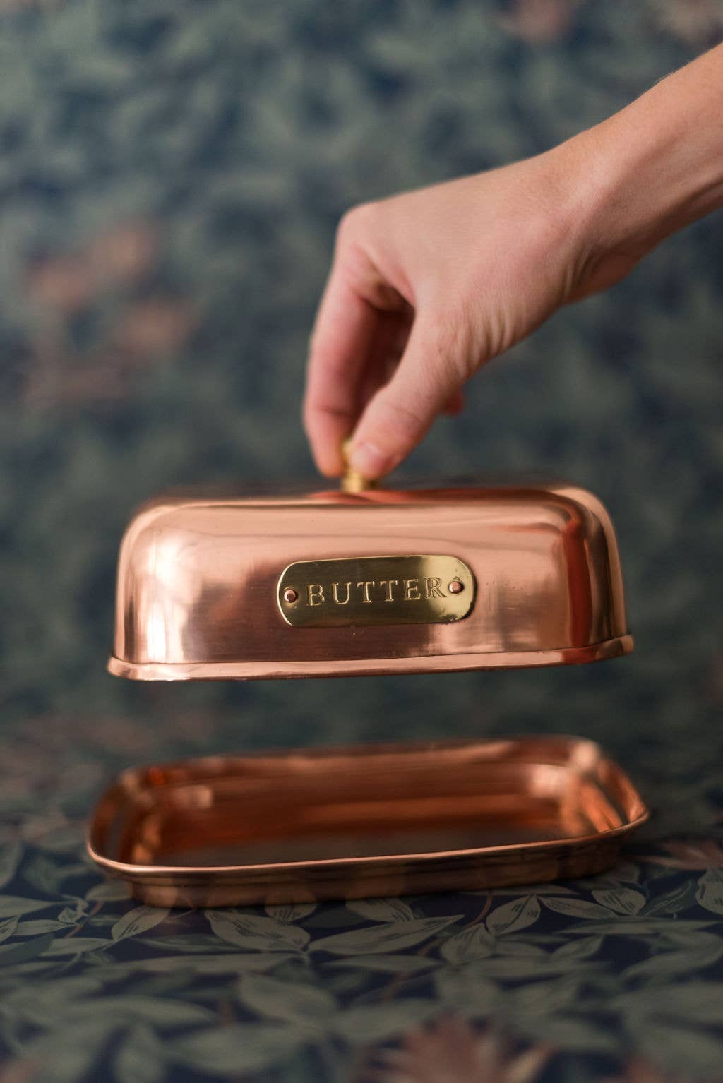 Copper butter dish with a hand lifting the lid on a textured surface