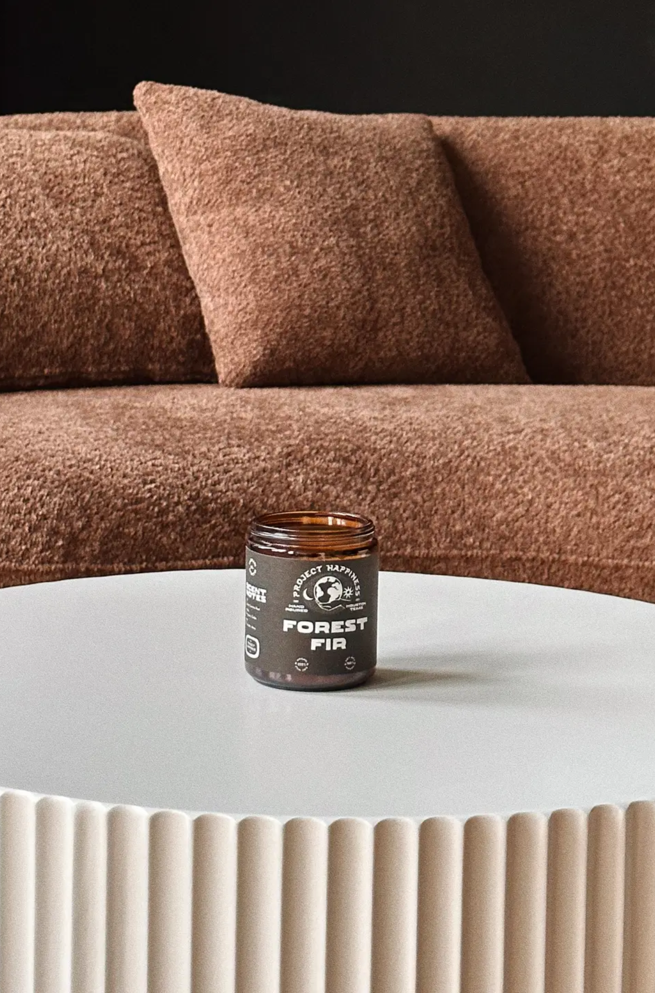 Project Happiness candle labeled 'Forest Fir' on a white table with a brown sofa in the background