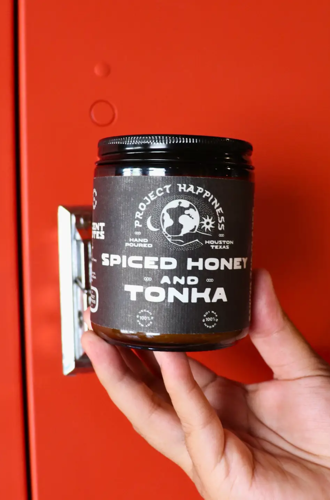 Hand holding a Project Happiness jar candle labeled 'Spiced Honey and Tonka' against a red background
